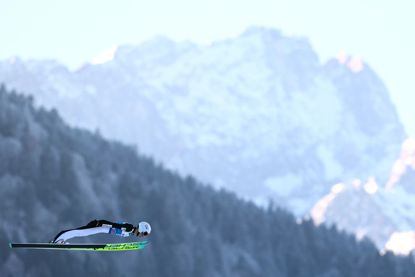 Kacper Tomasiak Zawodnik w trakcie lotu na nartach podczas konkursu skoków narciarskich, w tle ośnieżone góry oraz las.