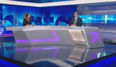 Kobieta i mężczyzna siedzą za stołem w studiu telewizyjnym, przed nimi mikrofony.