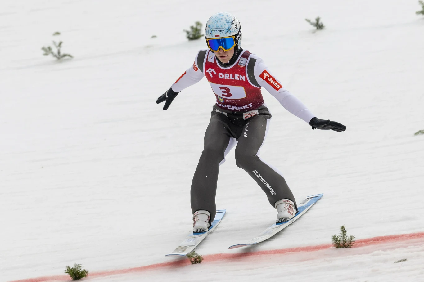 Zawodnik w kombinezonie narciarskim i kasku ląduje na nartach podczas konkursu skoków narciarskich, widoczna numeracja i sponsorzy na stroju, płaska, ośnieżona skocznia, kilka iglaków wystających z białej powierzchni.