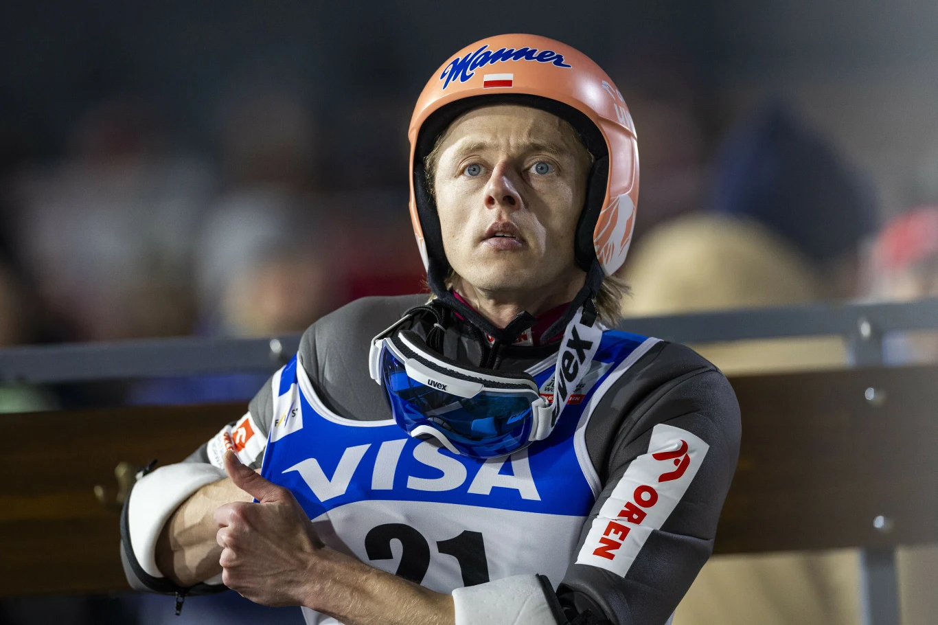 Skoczek narciarski w kombinezonie startowym, kasku i goglach trzyma ręce skrzyżowane na klatce piersiowej, z widocznym napięciem oczekuje na swoją kolej. W tle rozmyci kibice i elementy skoczni.