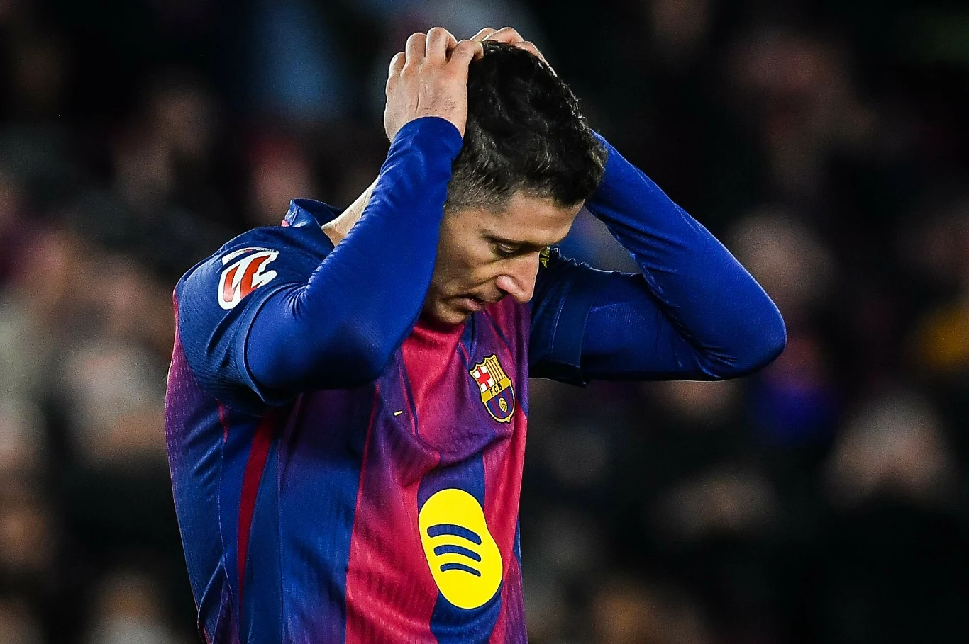 Piłkarz w stroju drużyny FC Barcelona trzyma ręce na głowie, wyrażając rozczarowanie lub frustrację. Jego twarz zwrócona jest w dół, a tło stanowią nieostre sylwetki publiczności.