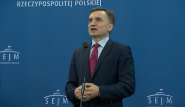 Mężczyzna w ciemnym garniturze i czerwonym krawacie przemawia do mikrofonu na tle niebieskiej ściany z napisem "RZECZYPOSPOLITA POLSKA" i logotypem Sejmu.