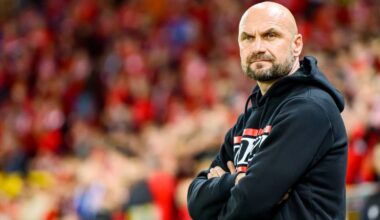 Łysy mężczyzna z brodą w czarnej bluzie z kapturem, stojący na tle kibic&oacute;w.