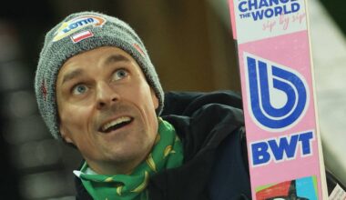 Skoczek narciarski w szarej czapce i ciemnej kurtce, trzymający różową nartę.