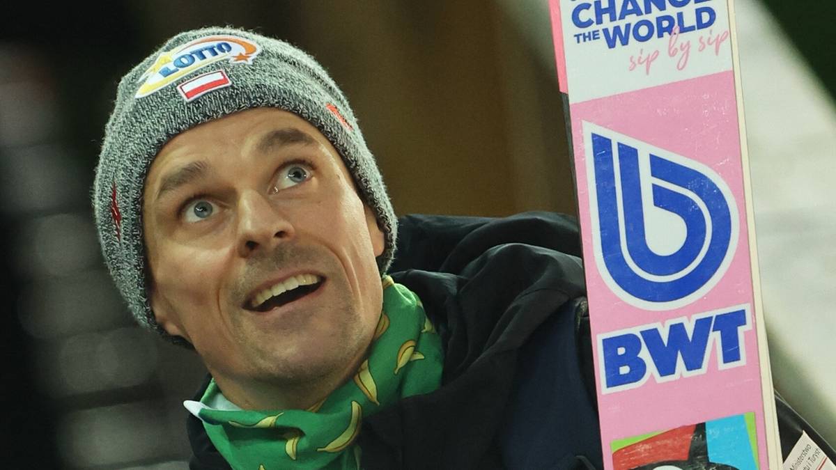 Skoczek narciarski w szarej czapce i ciemnej kurtce, trzymający różową nartę.