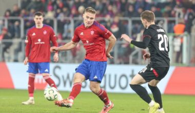 Piłkarz w czerwonej koszulce z niebieskimi spodenkami pr&oacute;buje odebrać piłkę zawodnikowi w czarnej koszulce.