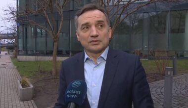 Mężczyzna w garniturze stoi przed budynkiem i m&oacute;wi do mikrofonu z logo Polsat News.