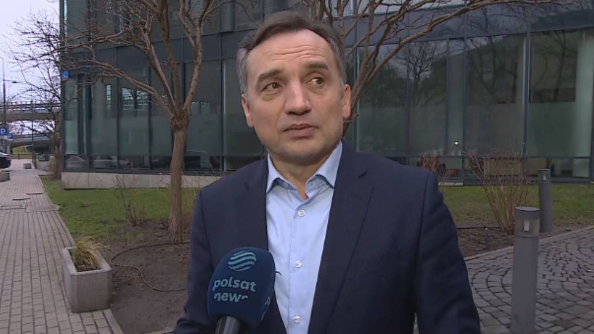 Mężczyzna w garniturze stoi przed budynkiem i m&oacute;wi do mikrofonu z logo Polsat News.