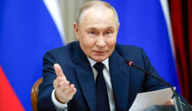 Vladimir Putin siedzi przy stole, gestykuluje prawą ręką i m&oacute;wi do mikrofonu. Na stole leży dokument. W tle widoczne są flagi Rosji.