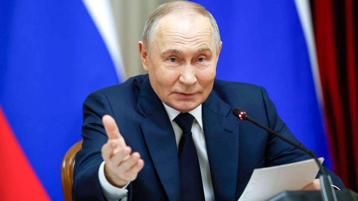 Vladimir Putin siedzi przy stole, gestykuluje prawą ręką i m&oacute;wi do mikrofonu. Na stole leży dokument. W tle widoczne są flagi Rosji.