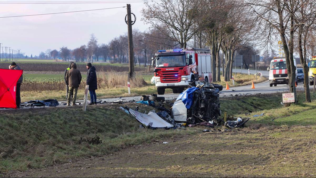 Miejsce wypadku drogowego z widocznym wrakiem samochodu, strażą pożarną i innymi pojazdami.