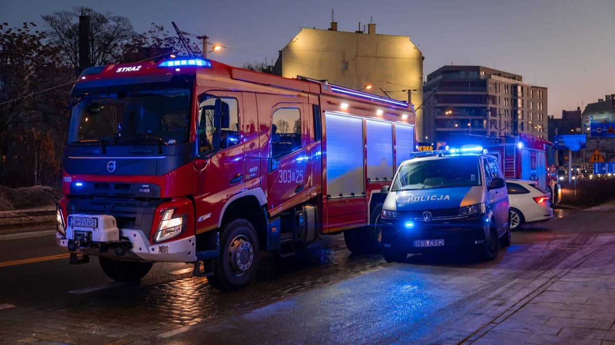 Samoch&oacute;d strażacki i radiow&oacute;z policyjny stojące na ulicy w nocy, oba pojazdy mają włączone niebieskie światła ostrzegawcze.