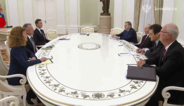 Grupa osób siedzi przy owalnym stole w eleganckim pomieszczeniu.