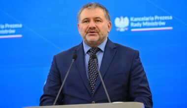 Mężczyzna w granatowym garniturze, białej koszuli i krawacie stoi przy m&oacute;wnicy, m&oacute;wiąc do mikrofonu. Tło jest niebieskie z napisem "Kancelaria Prezesa Rady Ministr&oacute;w".