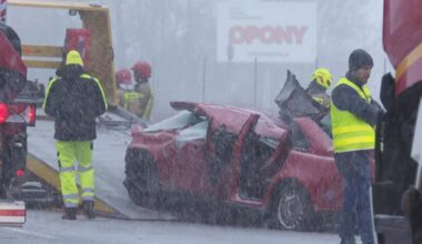 Uszkodzony czerwony samoch&oacute;d osobowy po wypadku drogowym, obok widoczni ratownicy w kamizelkach odblaskowych i ciężar&oacute;wka.