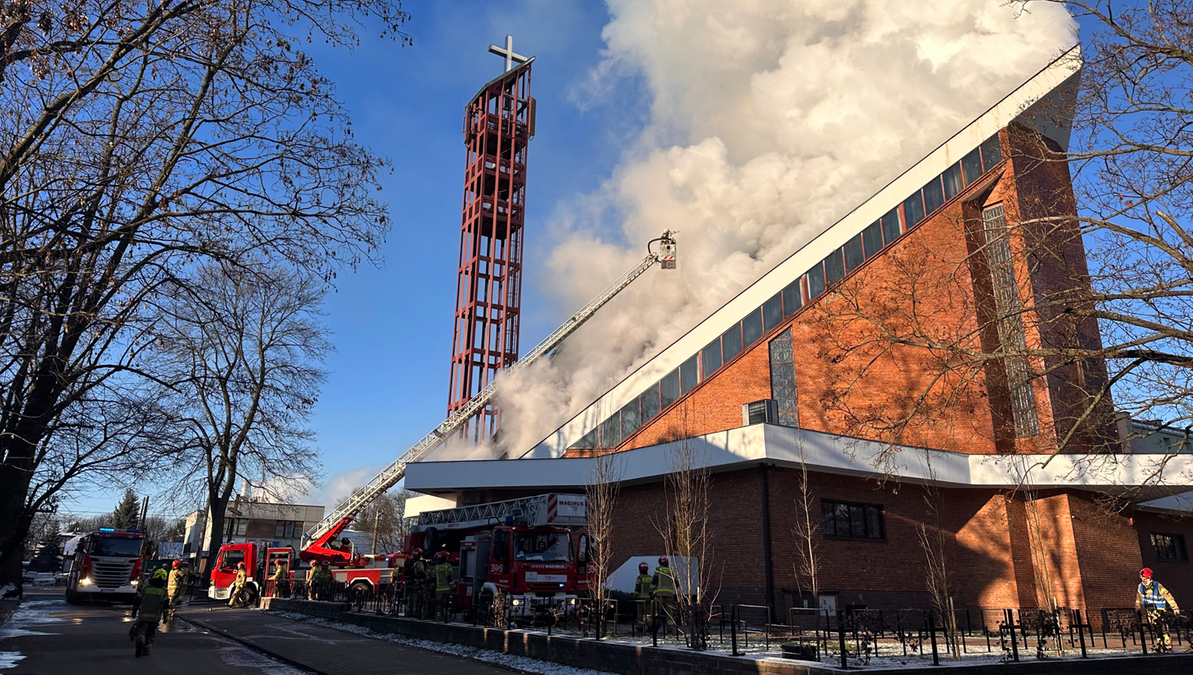Strażacy gaszą pożar kościoła, używając podnośnika i węży strażackich. Z dachu budowli wydobywają się kłęby dymu.
