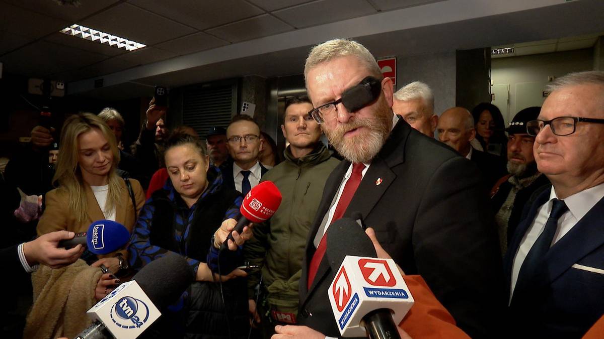 Mężczyzna z brodą i opaską na oku, ubrany w garnitur, przemawia do mikrofon&oacute;w.