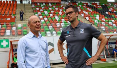 Dw&oacute;ch mężczyzn w strojach sportowych stoi na boisku piłkarskim przed trybunami.