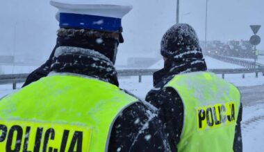 Dw&oacute;ch funkcjonariuszy policji w kamizelkach odblaskowych z napisem "POLICJA" stoi tyłem do widza na tle zaśnieżonej drogi.