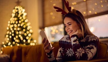 Kobieta w świątecznej opasce z rogami jelenia korzysta ze smartfona, siedząc na kanapie w tle widoczna jest choinka ozdobiona lampkami.