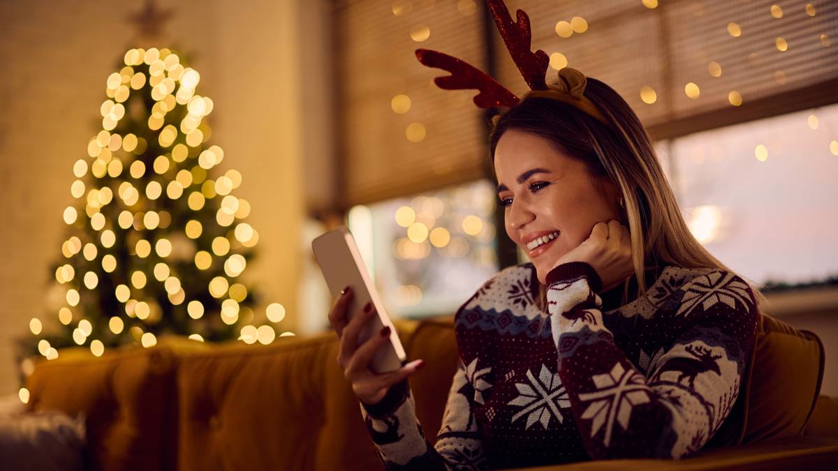 Kobieta w świątecznej opasce z rogami jelenia korzysta ze smartfona, siedząc na kanapie w tle widoczna jest choinka ozdobiona lampkami.