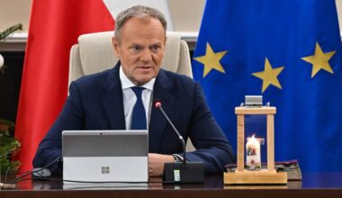 Donald Tusk siedzi przy stole, przed nim tablet i mikrofon, w tle flagi Polski i Unii Europejskiej.