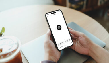 Ręce trzymają smartfon z wyraźnym logo ChatGPT na białym ekranie, nad zamkniętym laptopem na drewnianym stole, obok szklanki z mrożoną kawą i małej rośliny.