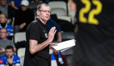 Trener siatk&oacute;wki w okularach i czarnej koszulce z notatkami w ręku, zwraca się do drużyny.