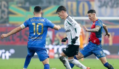 Piłkarze w białych i niebieskich strojach walczą o piłkę na boisku.