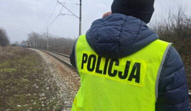 Policjant w kamizelce odblaskowej z napisem "POLICJA" na plecach stoi przy torach kolejowych.