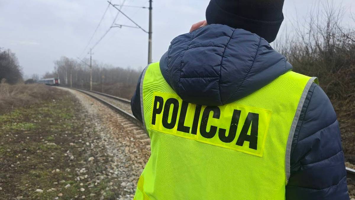 Policjant w kamizelce odblaskowej z napisem "POLICJA" na plecach stoi przy torach kolejowych.