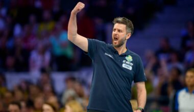 Trener piłki ręcznej w ciemnej koszulce z podniesioną prawą ręką, krzyczący z radości lub motywacji na tle trybun kibic&oacute;w.