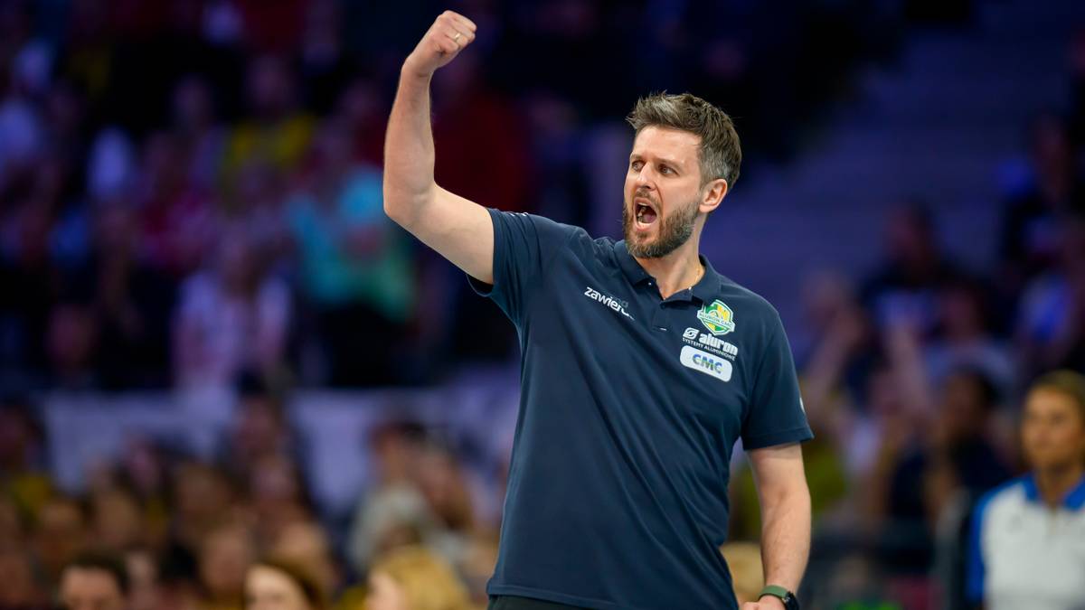 Trener piłki ręcznej w ciemnej koszulce z podniesioną prawą ręką, krzyczący z radości lub motywacji na tle trybun kibic&oacute;w.