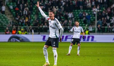 Piłkarz w białej koszulce z czarnymi spodenkami, z uniesioną prawą ręką, na boisku piłkarskim.