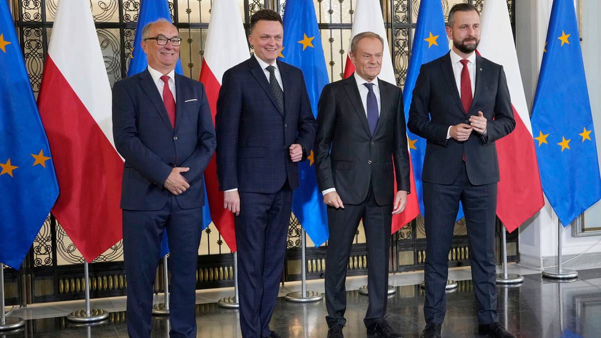 Czterech mężczyzn w garniturach, w tym Donald Tusk, Władysław Kosiniak-Kamysz i Szymon Hołownia, stoi przed rzędem flag.