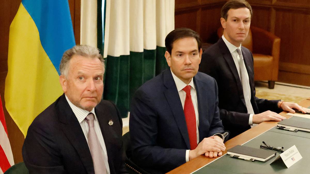 Trzech mężczyzn siedzi przy stole konferencyjnym. Na pierwszym planie po lewej znajduje się mężczyzna z siwymi włosami i jasnym garniturze. W środku siedzi mężczyzna o ciemnych włosach, w ciemnym garniturze i czerwonym krawacie. Po prawej stoi wysoki, szczupły mężczyzna w ciemnym garniturze i jasnym krawacie. Za nimi widoczne są flagi Ukrainy i USA.