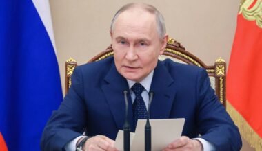 Władimir Putin siedzi przy stole, trzymając w dłoniach kartki z tekstem i m&oacute;wiąc do mikrofon&oacute;w.