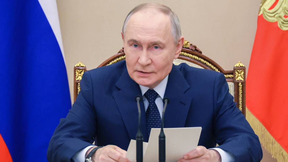 Władimir Putin siedzi przy stole, trzymając w dłoniach kartki z tekstem i m&oacute;wiąc do mikrofon&oacute;w.