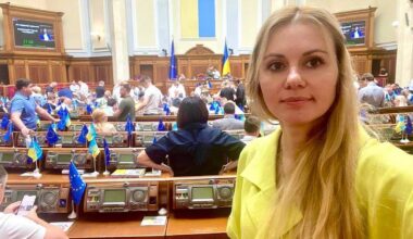 Anna Skorokhod, ukraińska parlamentarzystka, stoi przed rzędem miejsc w sali posiedzeń parlamentu, z innymi posłami widocznymi w tle.