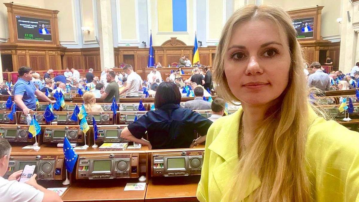 Anna Skorokhod, ukraińska parlamentarzystka, stoi przed rzędem miejsc w sali posiedzeń parlamentu, z innymi posłami widocznymi w tle.