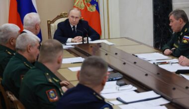 Władimir Putin siedzi przy stole konferencyjnym, rozmawiając z grupą rosyjskich dow&oacute;dc&oacute;w wojskowych.