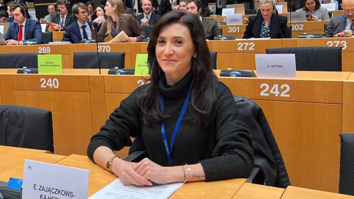 Kobieta siedzi przy stole w sali konferencyjnej, przed nią leżą dokumenty i identyfikator z napisem "EWA ZAJĄCZKOWSKA-HERNIK"