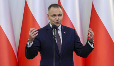 Mężczyzna w garniturze przemawia przy m&oacute;wnicy na tle flag Polski.