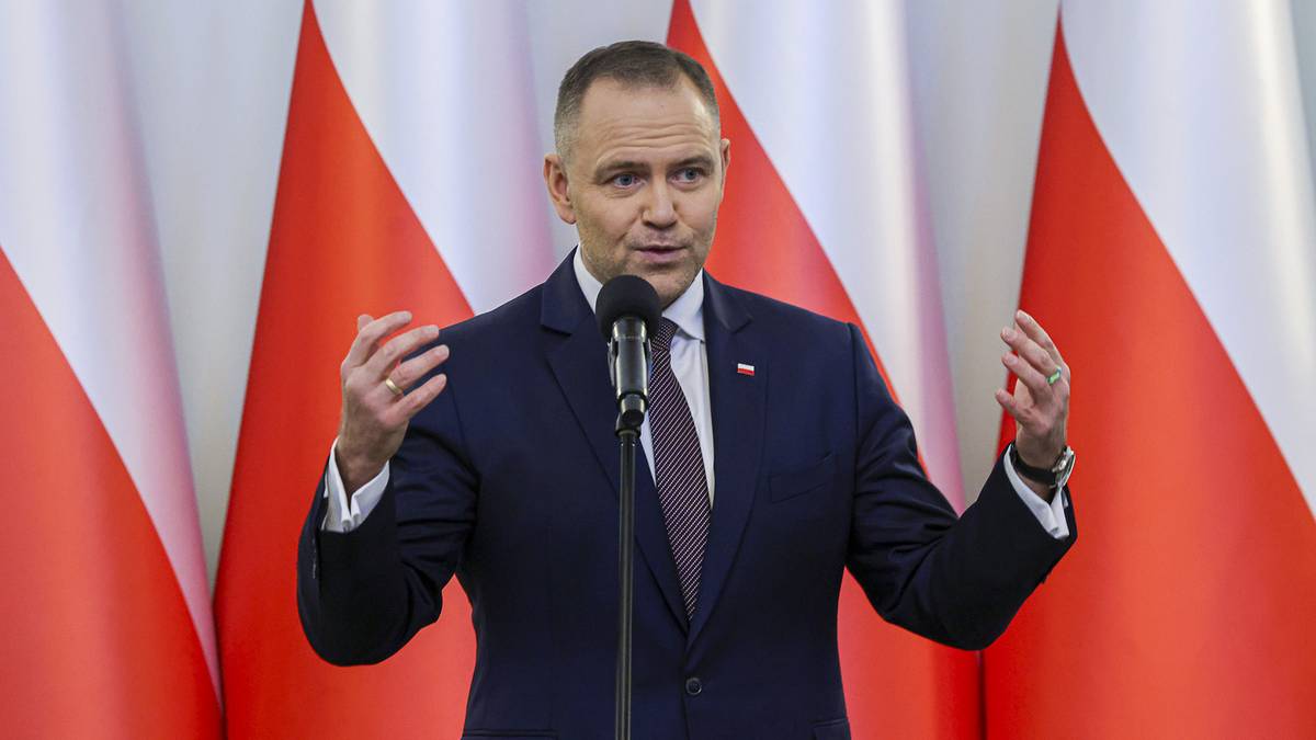 Mężczyzna w garniturze przemawia przy m&oacute;wnicy na tle flag Polski.