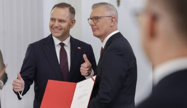 Dw&oacute;ch mężczyzn w garniturach, jeden z nich uśmiecha się i pokazuje kciuk w g&oacute;rę, trzymając czerwony folder.