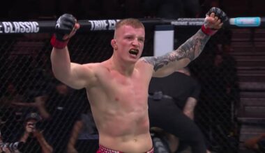 Młody mężczyzna z krótkimi blond włosami, wytatuowanymi ramionami i opaskami na nadgarstkach, w okrągłym ringu, z rękami uniesionymi w geście triumfu, krzyczy.