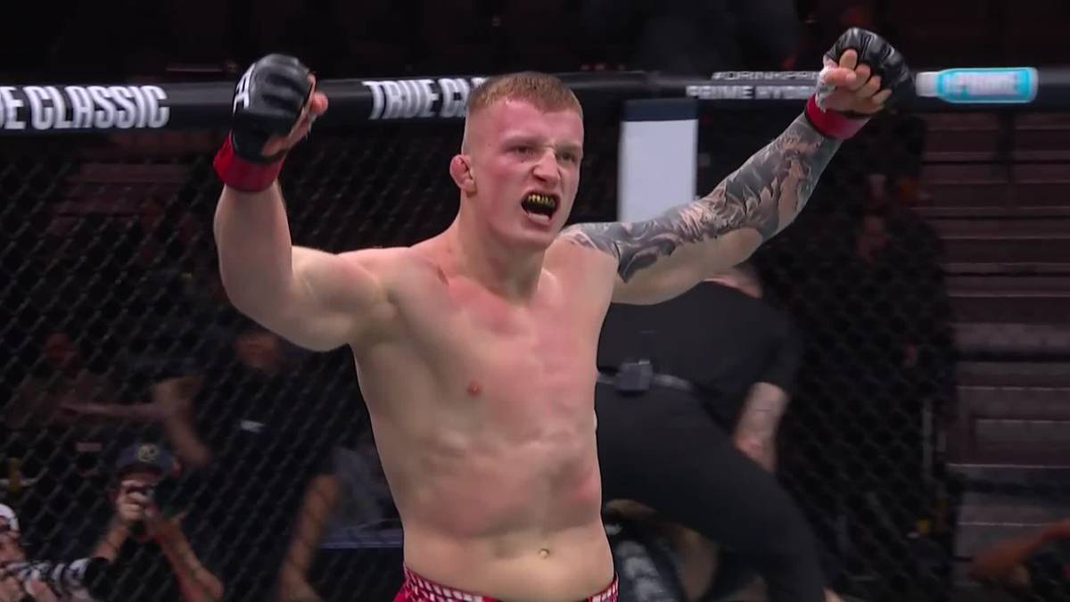 Młody mężczyzna z krótkimi blond włosami, wytatuowanymi ramionami i opaskami na nadgarstkach, w okrągłym ringu, z rękami uniesionymi w geście triumfu, krzyczy.