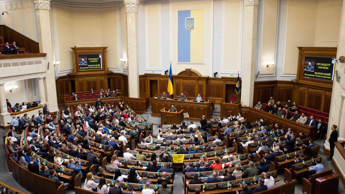 Widok na salę obrad parlamentu Ukrainy z widownią siedzącą na p&oacute;łkolistych ławach, z m&oacute;wnicą na czele i flagami państwowymi.