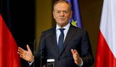 Donald Tusk w garniturze stoi przy mównicy z mikrofonami, gestykulując rękami. W tle widoczne flagi Polski i Niemiec.
