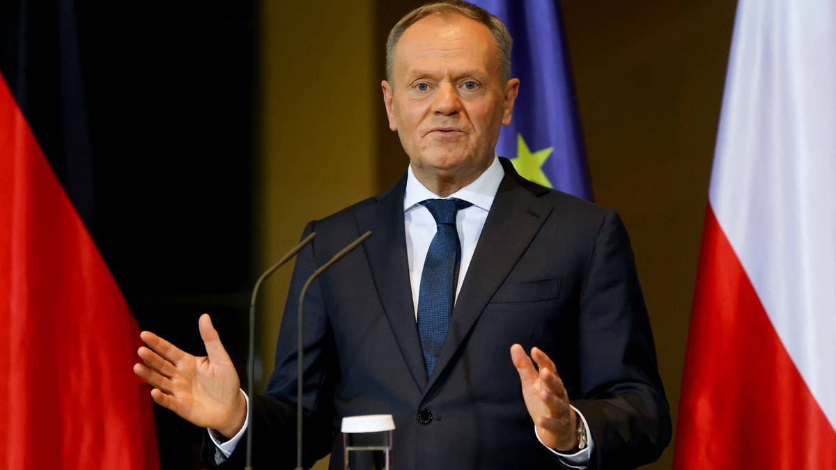 Donald Tusk w garniturze stoi przy mównicy z mikrofonami, gestykulując rękami. W tle widoczne flagi Polski i Niemiec.